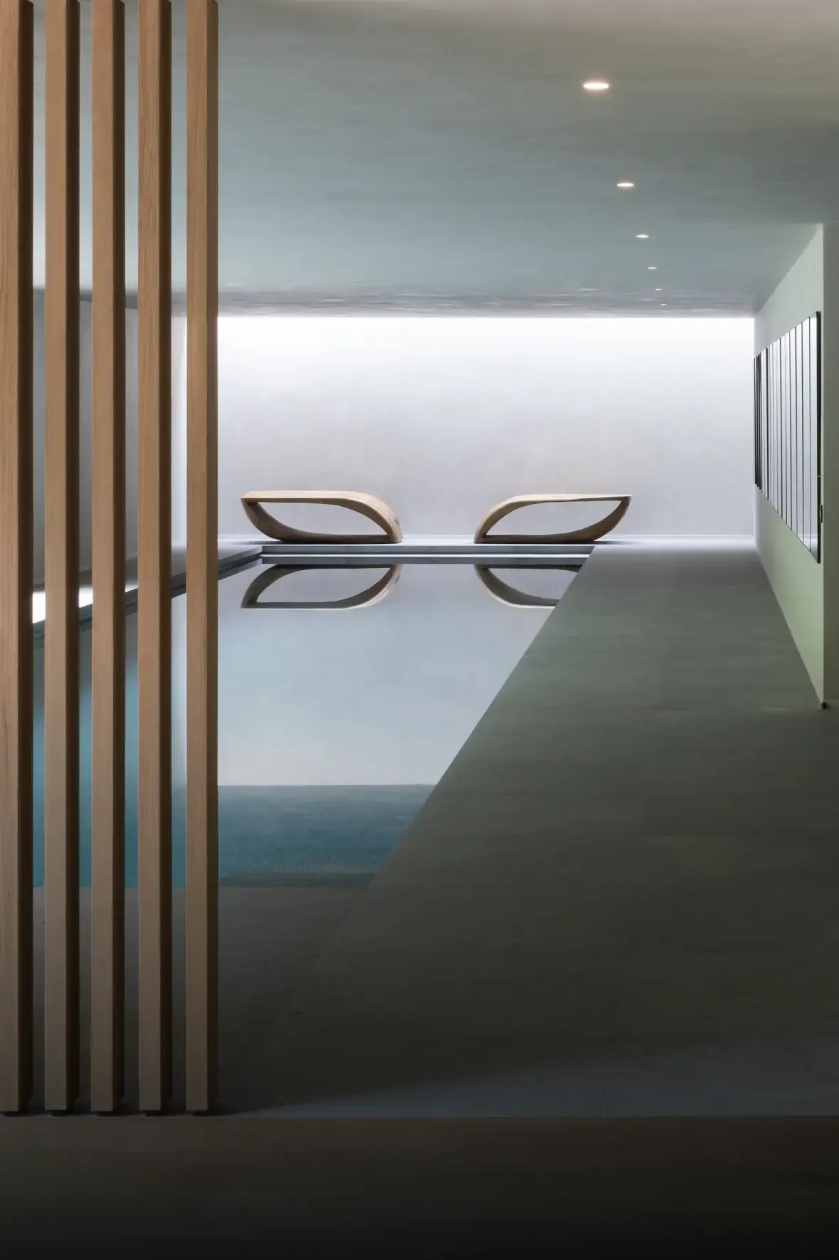 Modern indoor pool area with a serene ambiance. Wooden slats on the left, sleek pool in the center, and two curved loungers at the far end.