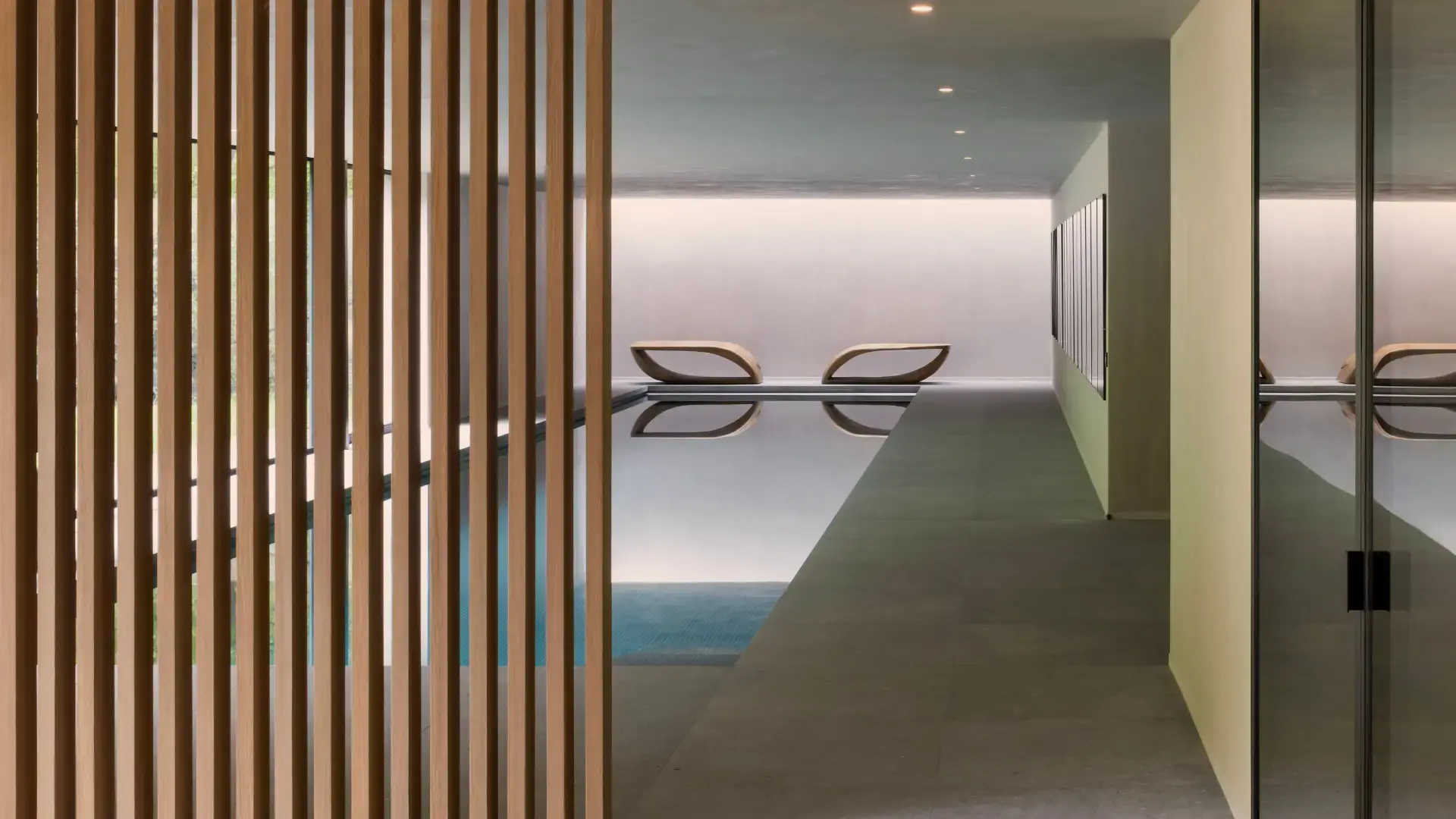 Modern indoor pool area with a serene ambiance. Wooden slats on the left, sleek pool in the center, and two curved loungers at the far end.