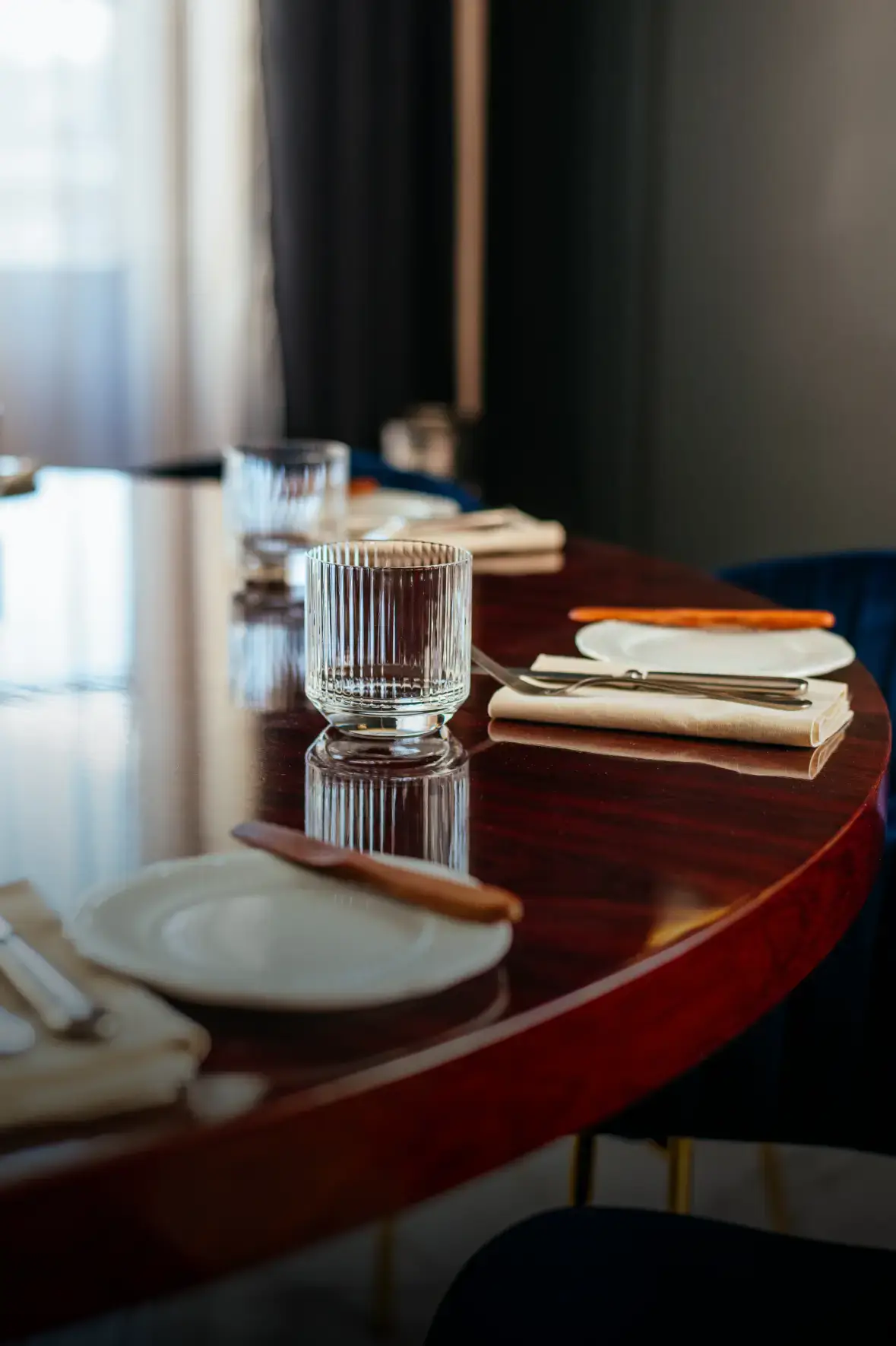 A polished wooden table is set with elegant, empty glasses, white plates, and cream napkins. The mood is formal and inviting, with soft lighting.