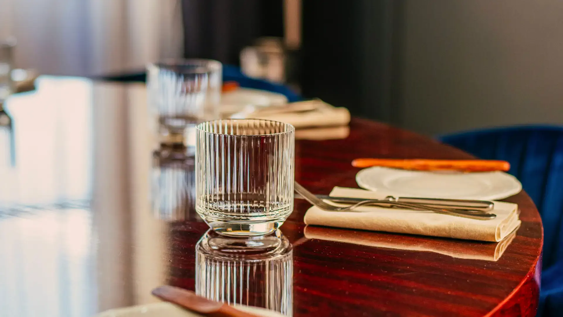 A polished wooden table is set with elegant, empty glasses, white plates, and cream napkins. The mood is formal and inviting, with soft lighting.