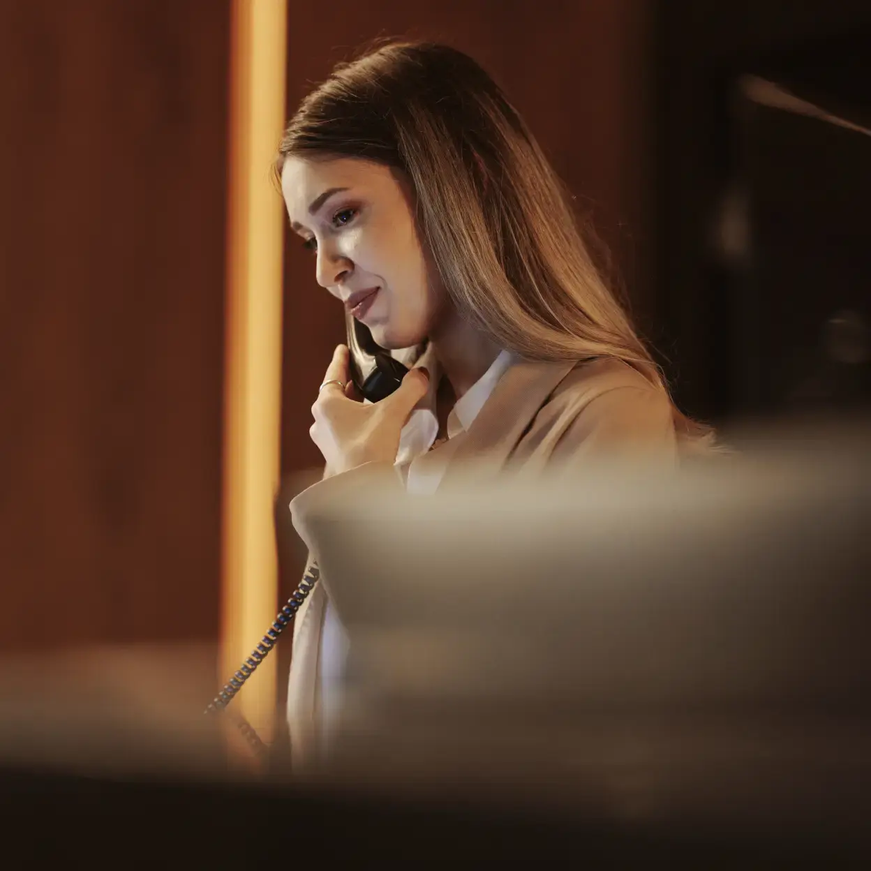 Woman in beige blazer smiling while talking on the phone in an office.