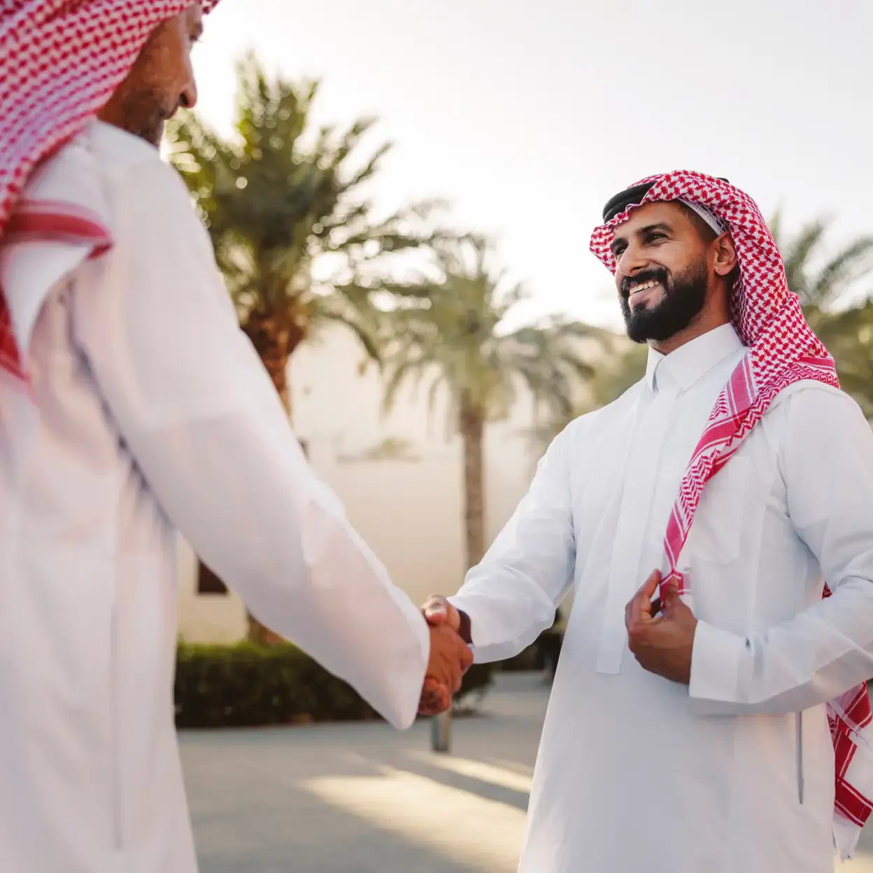 Two men in traditional Middle Eastern attire shake hands outdoors, smiling warmly. Palm trees and a bright sky create a friendly, welcoming atmosphere.