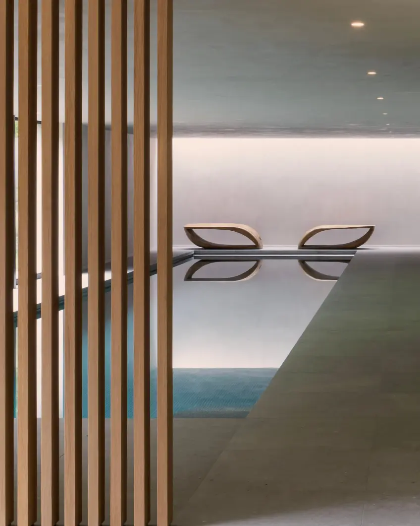 Modern indoor pool with wooden slats on the left, creating a minimalistic look. Two stylish lounge chairs reflect calmly in the water, exuding tranquility.