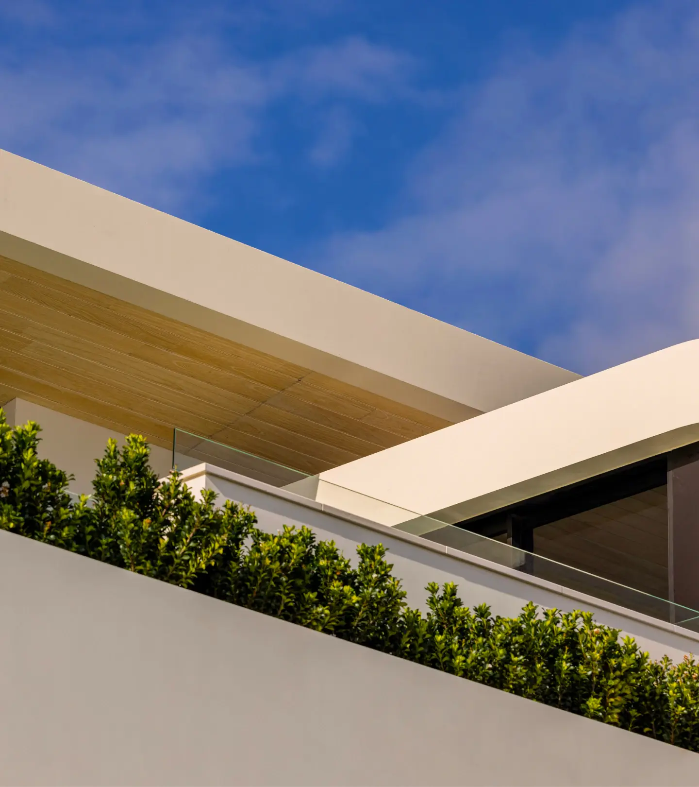 Modern architectural facade with sleek lines, glass railings, and green shrubs, set against a clear blue sky; conveys elegance and tranquility.
