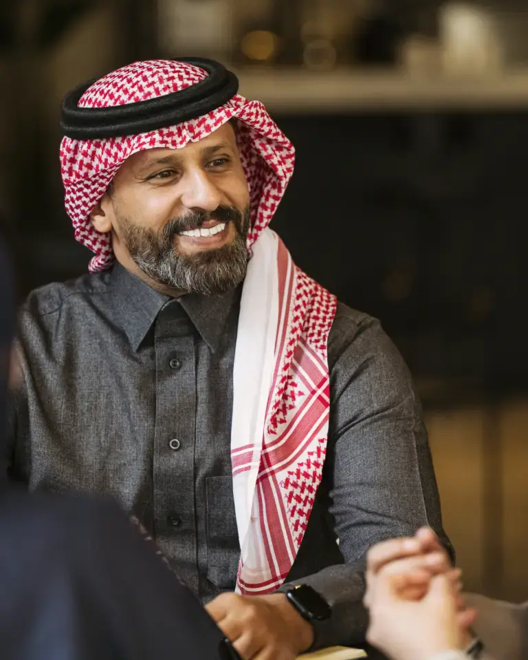 Smiling man in traditional Middle Eastern attire, including a red and white checkered keffiyeh and dark shirt, engaging in conversation in a warm setting.