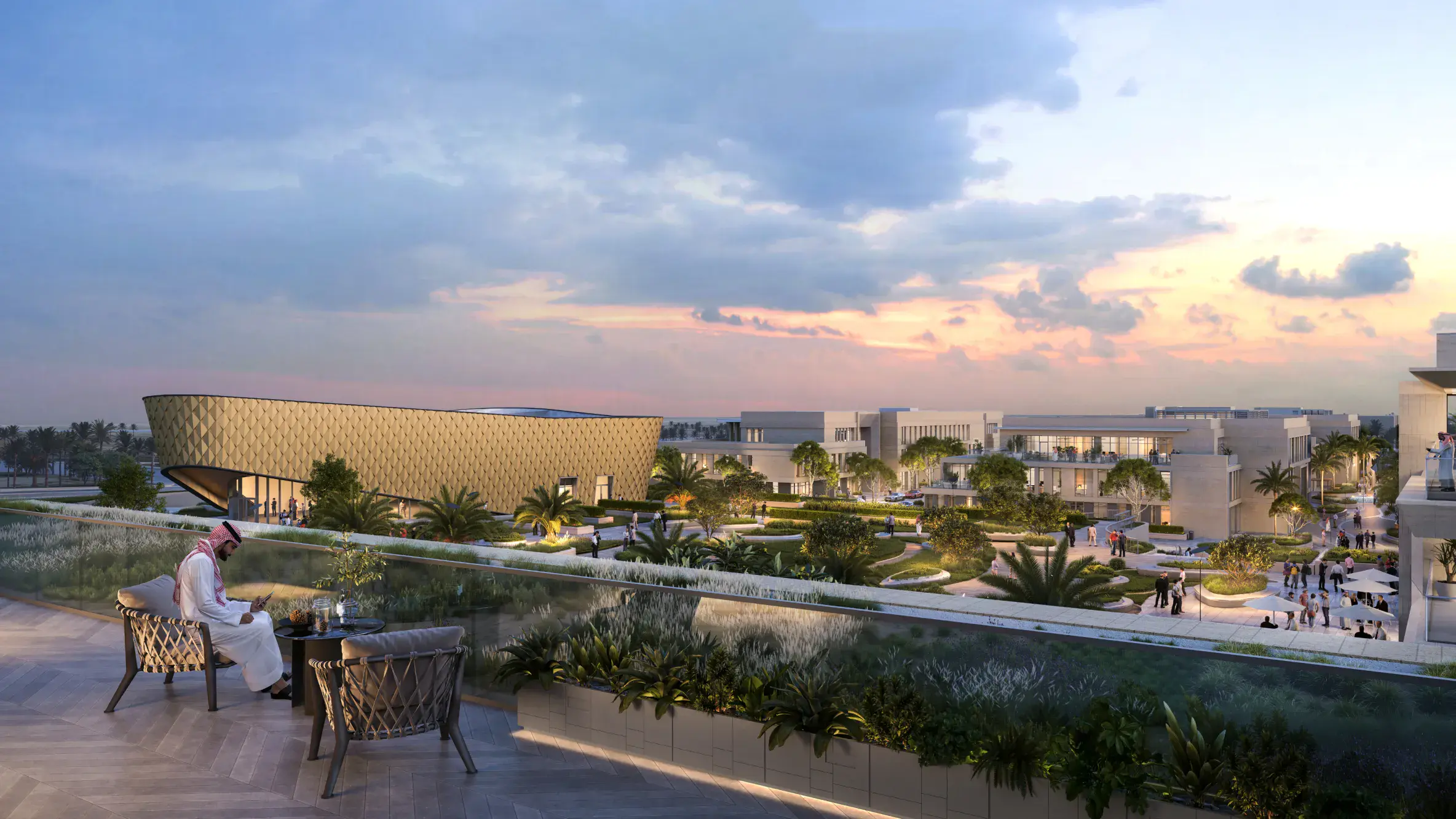 A person in traditional attire sits on a terrace overlooking a modern architectural complex at sunset. Lush greenery and a serene sky enhance the tranquil scene.