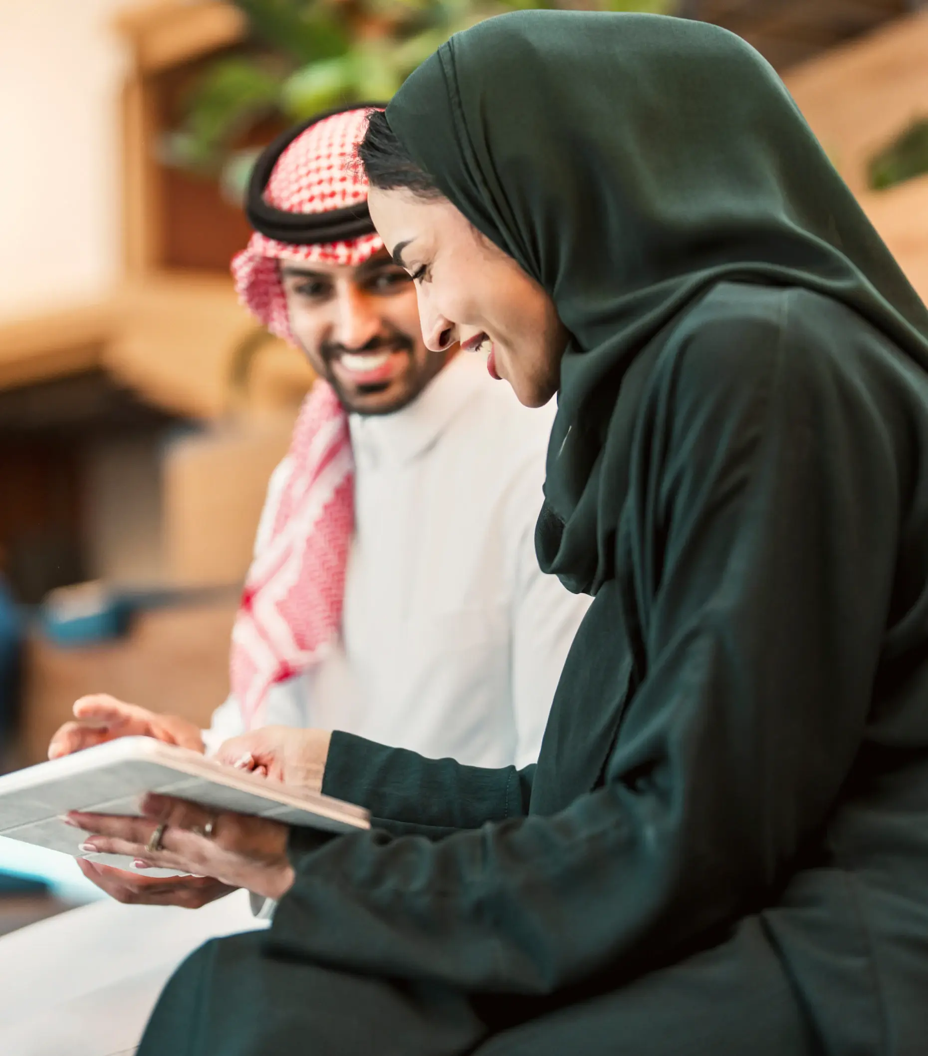 A man and woman, both in traditional Middle Eastern attire, smile while looking at a tablet together. They appear engaged and happy in a well-lit setting.
