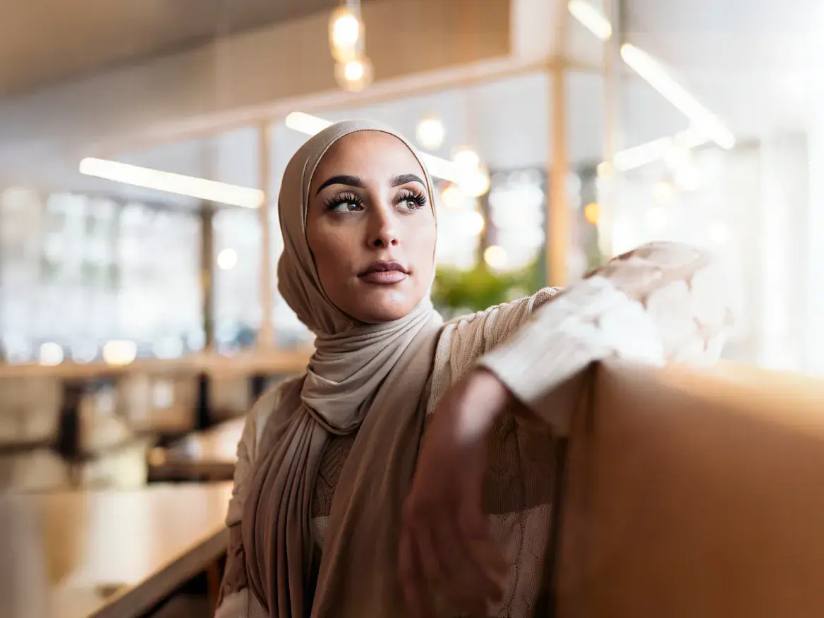 Saudi businesswoman in modern conservative clothing sitting indoors, gazing thoughtfully into the distance.