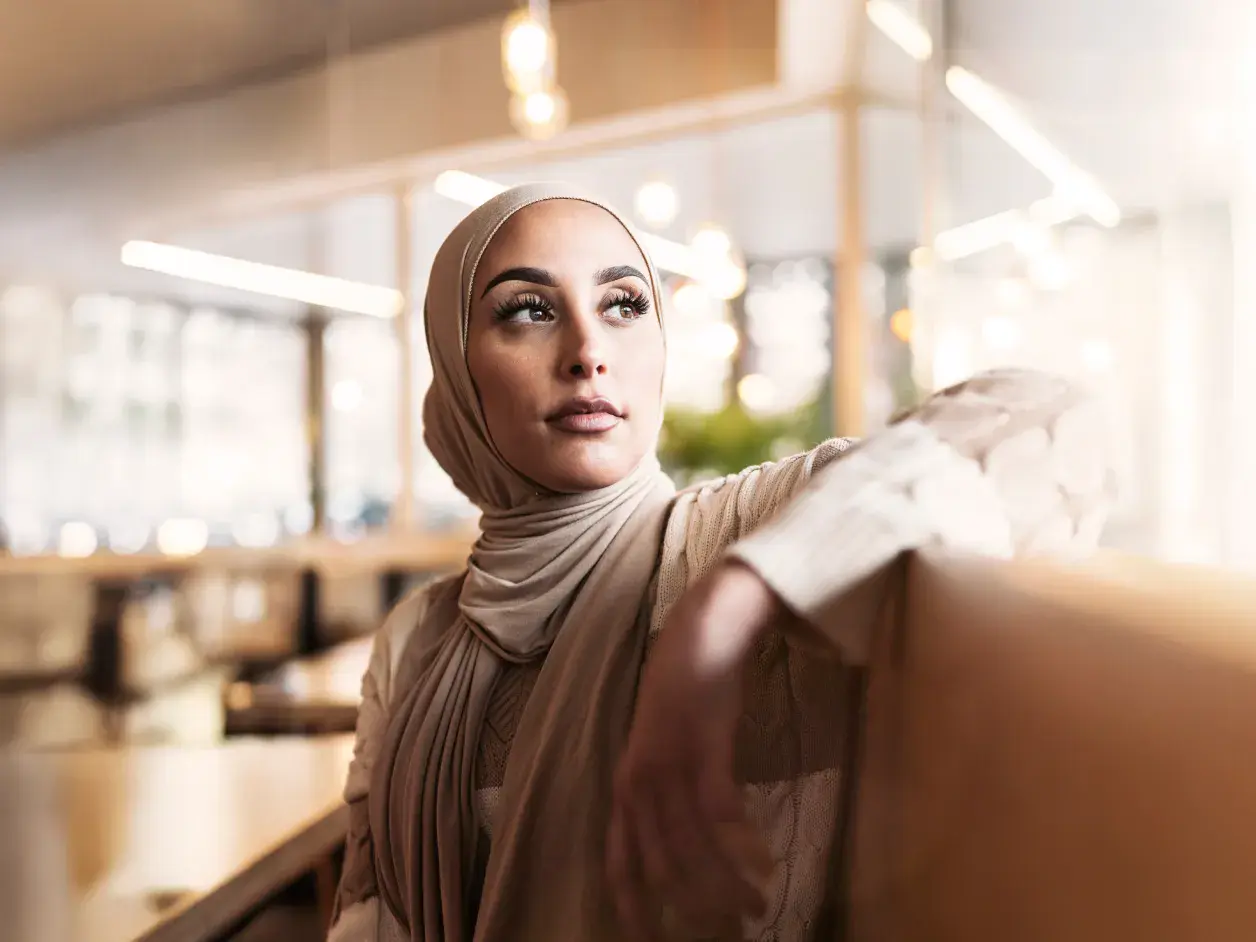 Saudi businesswoman in modern conservative clothing sitting indoors, gazing thoughtfully into the distance.