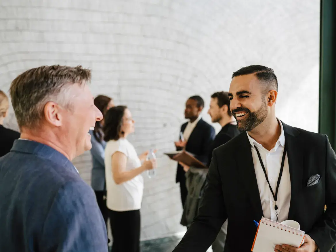 Saudi businessman in modern professional attire greeting a Western businessman, with networking in the background.