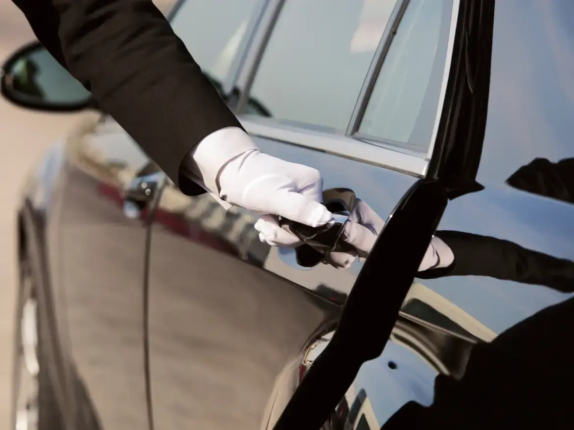 A gloved hand opens a sleek black car door, reflecting sunlight. The formal attire suggests a professional chauffeur, conveying luxury and service.