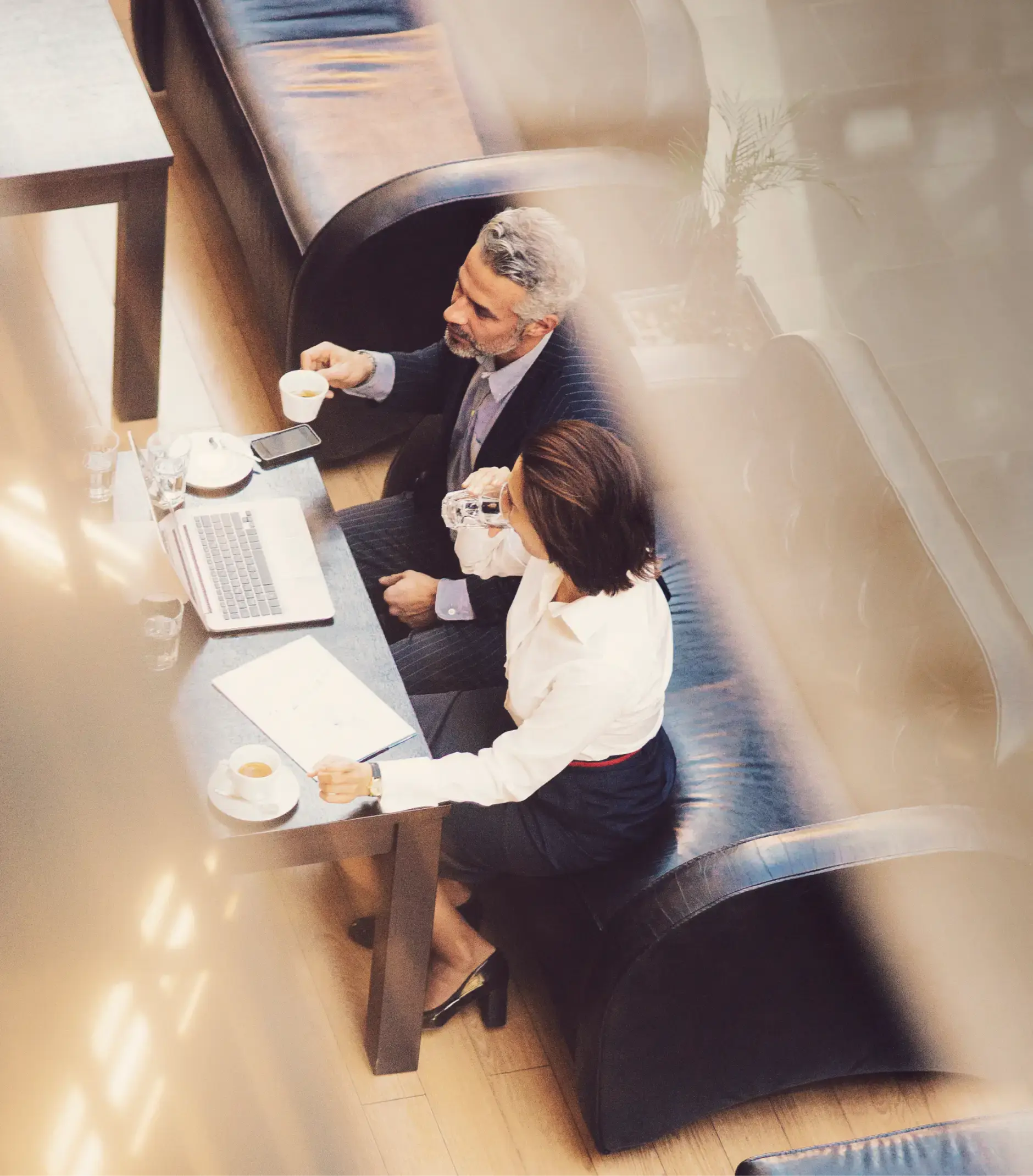 Two professionals drinking coffee and water while working on a laptop at a cafe table with documents and smartphones.