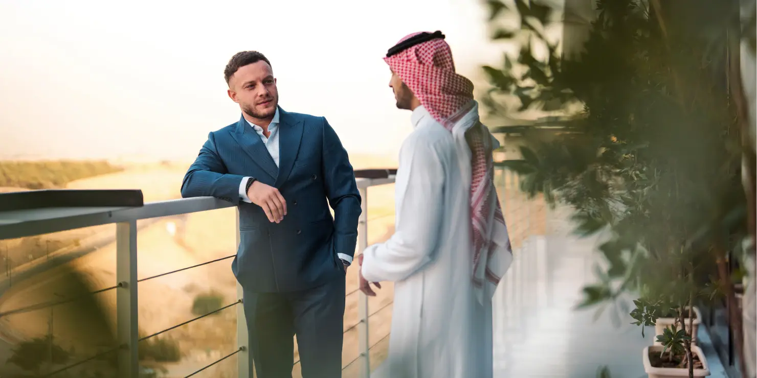 Two men converse on a sunny balcony, one in a blue suit, the other in traditional Middle Eastern attire. 