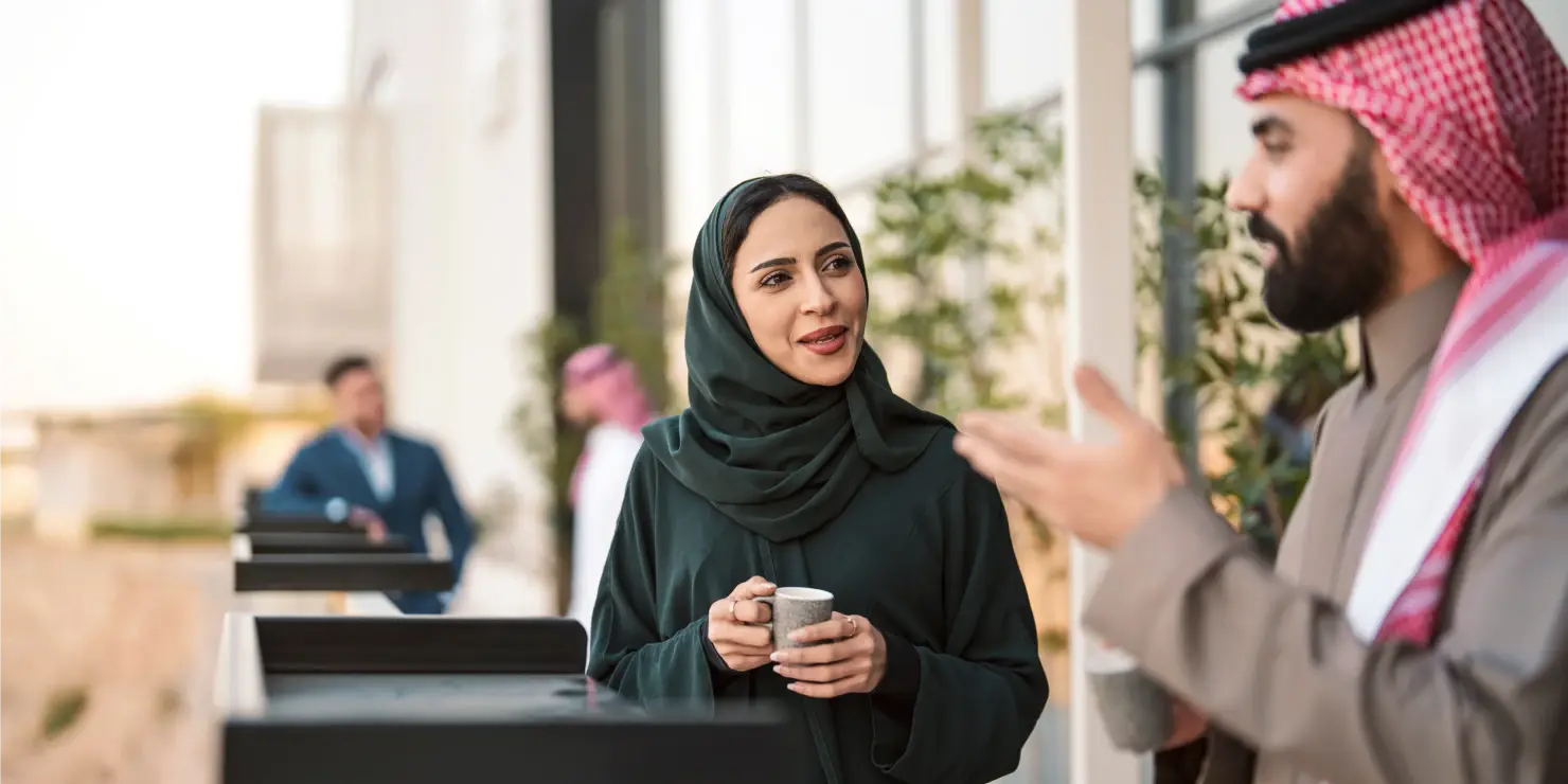 A woman in a black hijab and a man in traditional attire converse over coffee outdoors, smiling. Two men in the background also enjoy their drinks.
