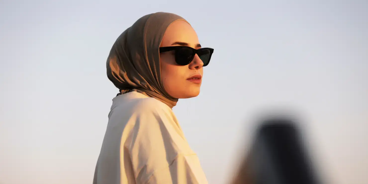 Woman wearing a brown hijab and sunglasses, dressed in a light-colored garment, standing outdoors against a clear sky background.